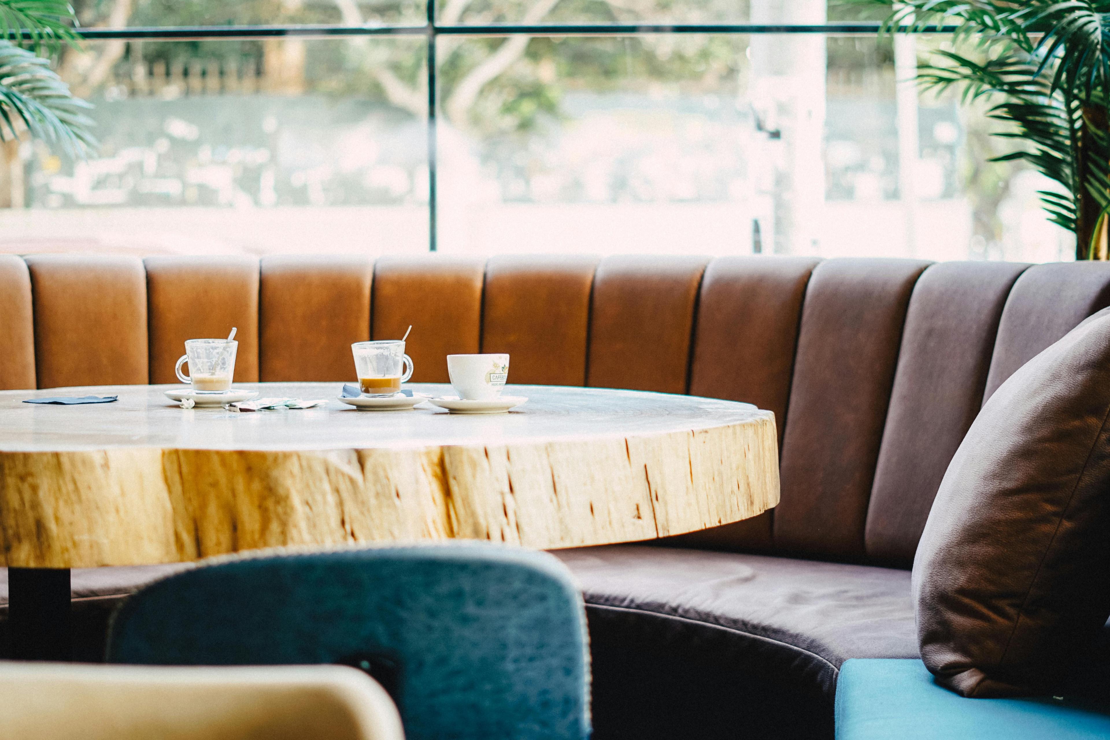 Café-Lounge mit rundem Massivholztisch, auf dem drei Kaffeetassen stehen, dahinter geschwungene Polsterbank in Braun-Grau-Tönen vor großer Glasfront mit Grünpflanzen.