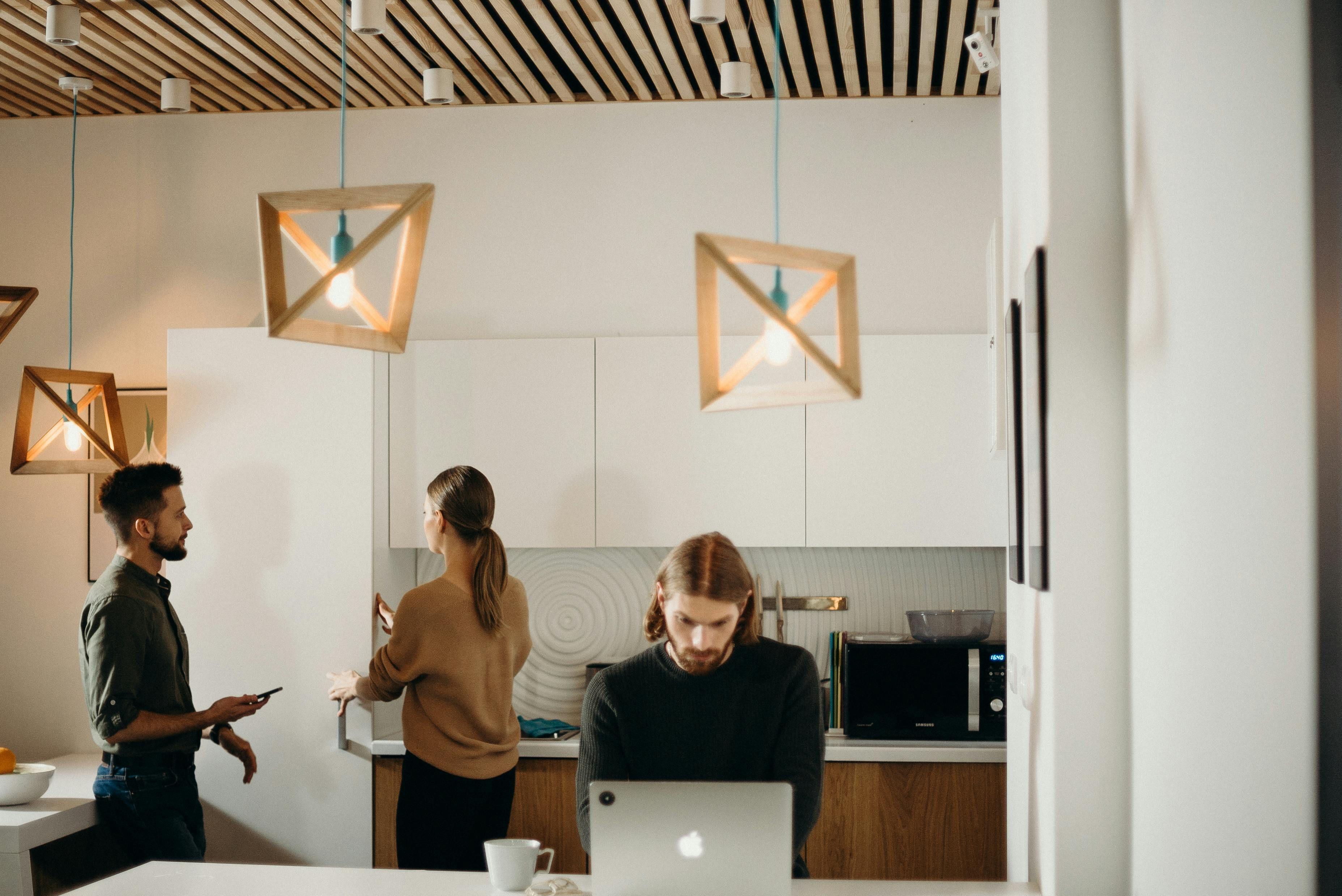 Moderne Büroküche mit drei Personen, geometrischen Holz-Pendelleuchten und Lamellendecke, im Vordergrund Person am Laptop, zwei Kollegen im Gespräch im Hintergrund.