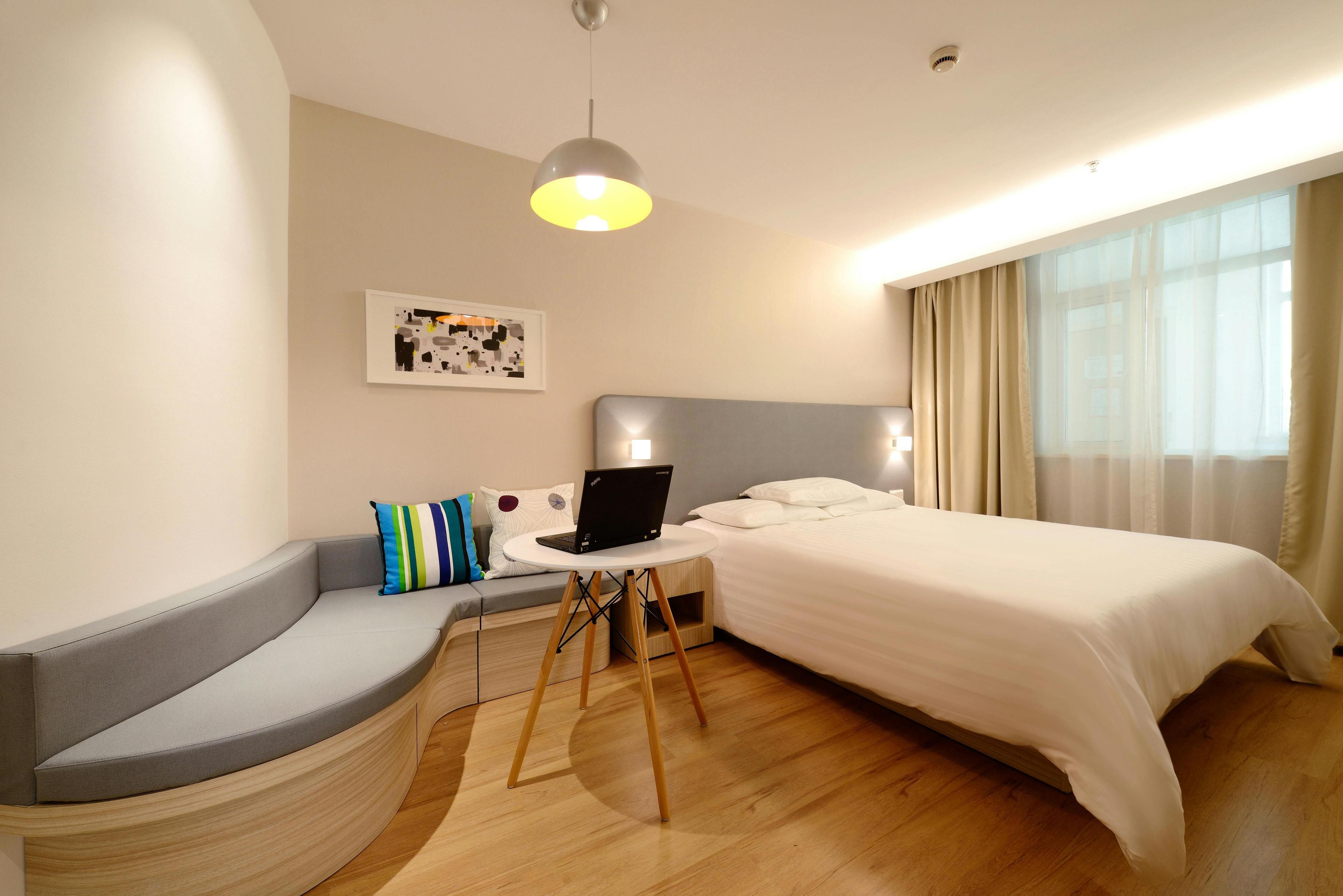 Modernes Hotelzimmer mit Doppelbett, grauer Sitzbank, rundem Arbeitstisch und hellem Holzboden, beleuchtet durch große Fensterfront mit beigen Vorhängen.