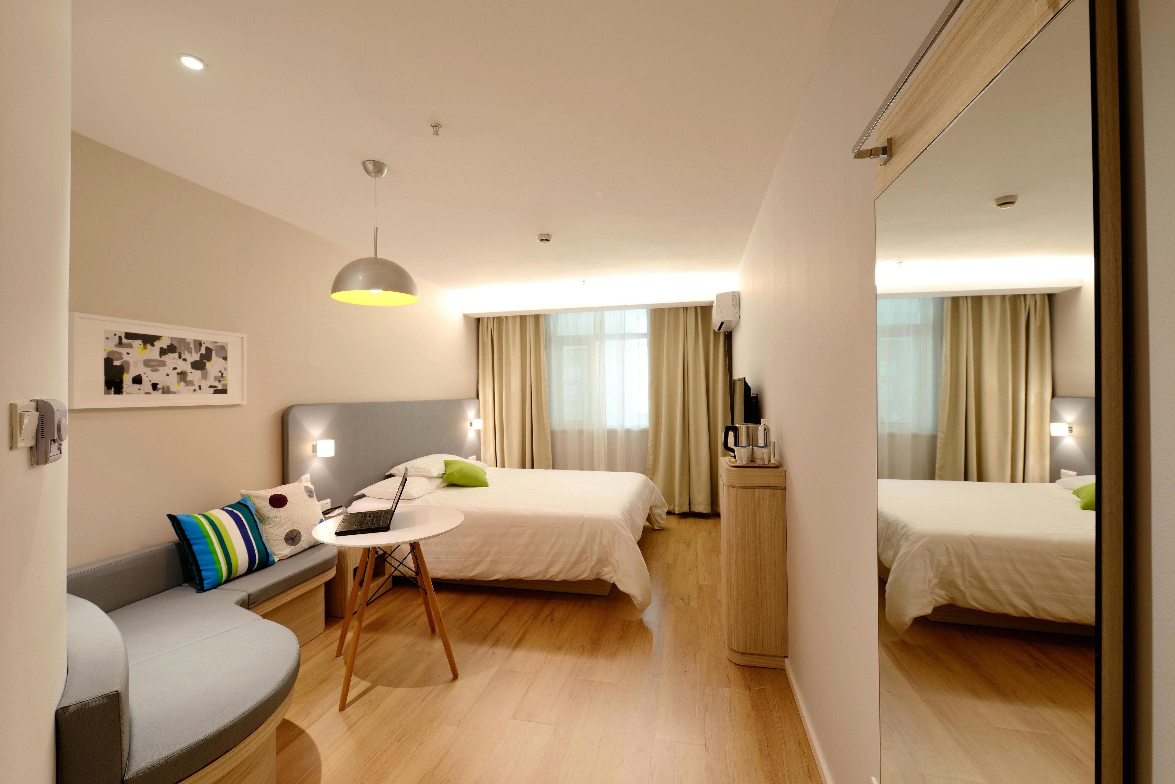 Helles Hotelzimmer mit Doppelbett, grauem Sofa, Spiegelschrank und rundem Arbeitstisch, Holzboden und großes Fenster mit beigen Vorhängen.