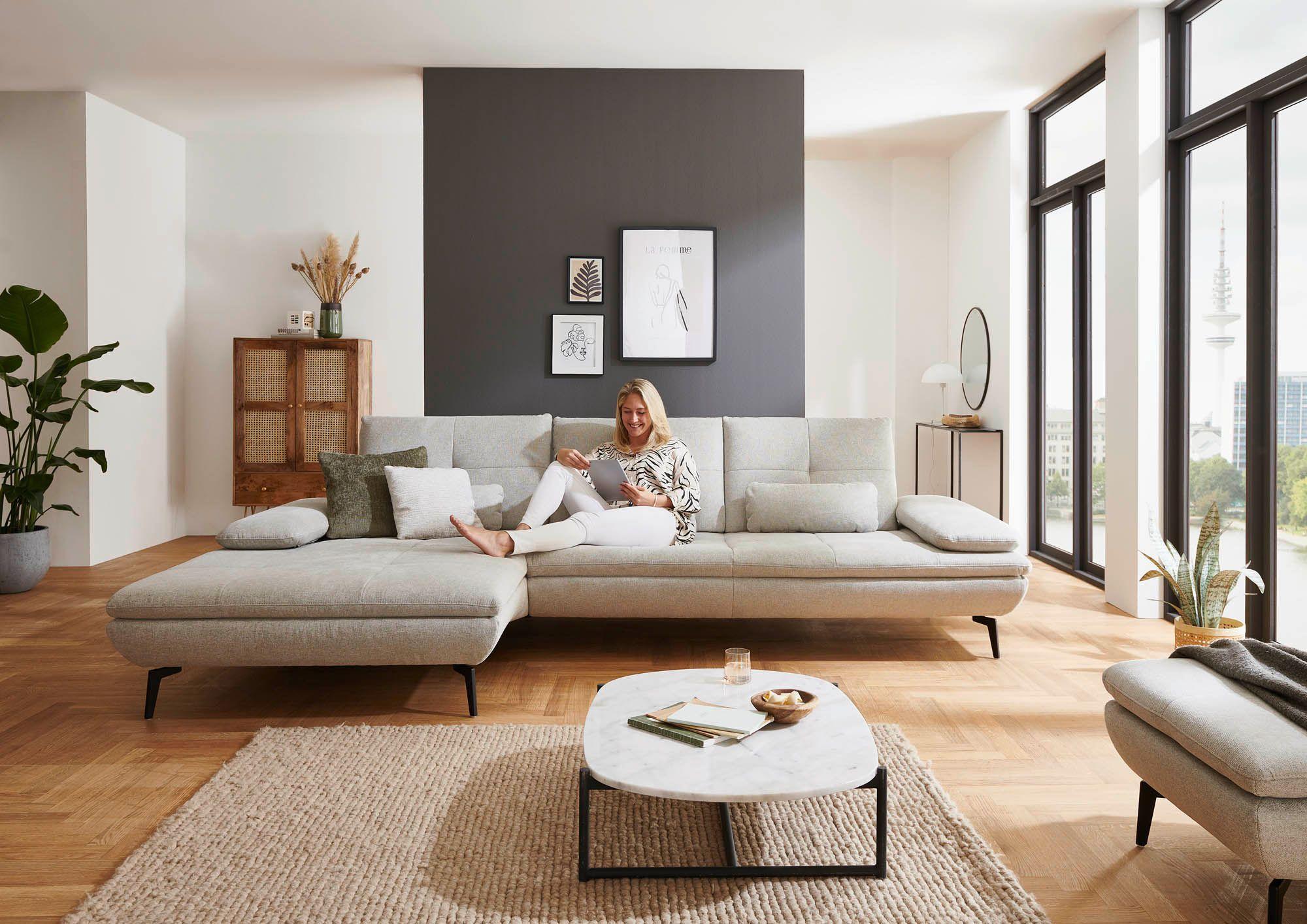 Sofa in ecrufarbenem Stoff mit Longchair links und Armlehne rechts, Füße Metall schwarz, auf welcher eine Frau sitzt und liest in der Mitte des Sofas