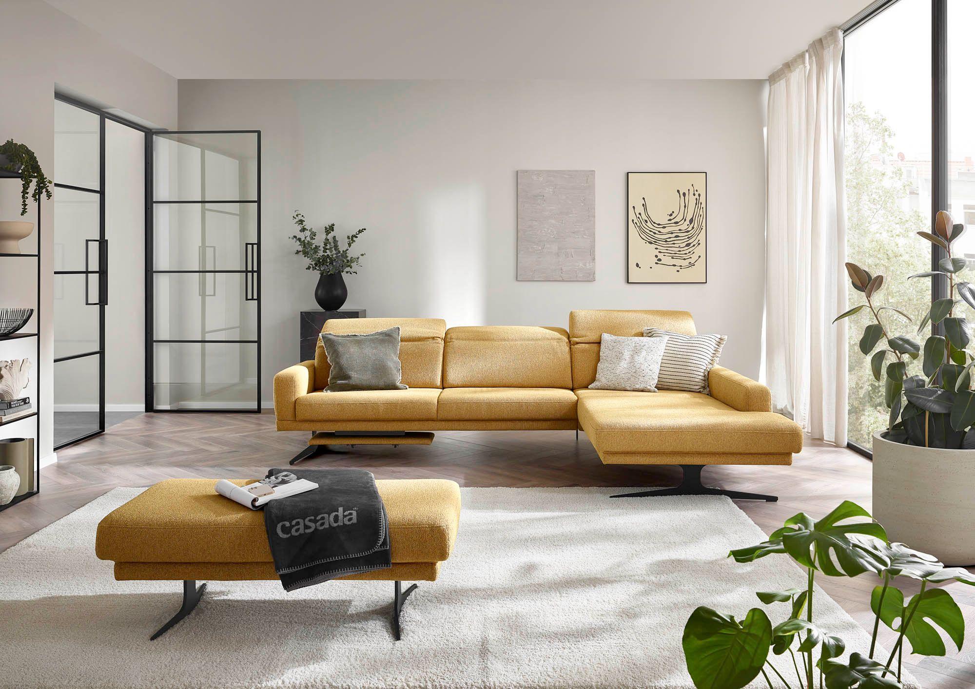 Sofa in patchoulifarbenem Stoff mit Armlehne links und Longchair rechts, davor steht ein Hocker