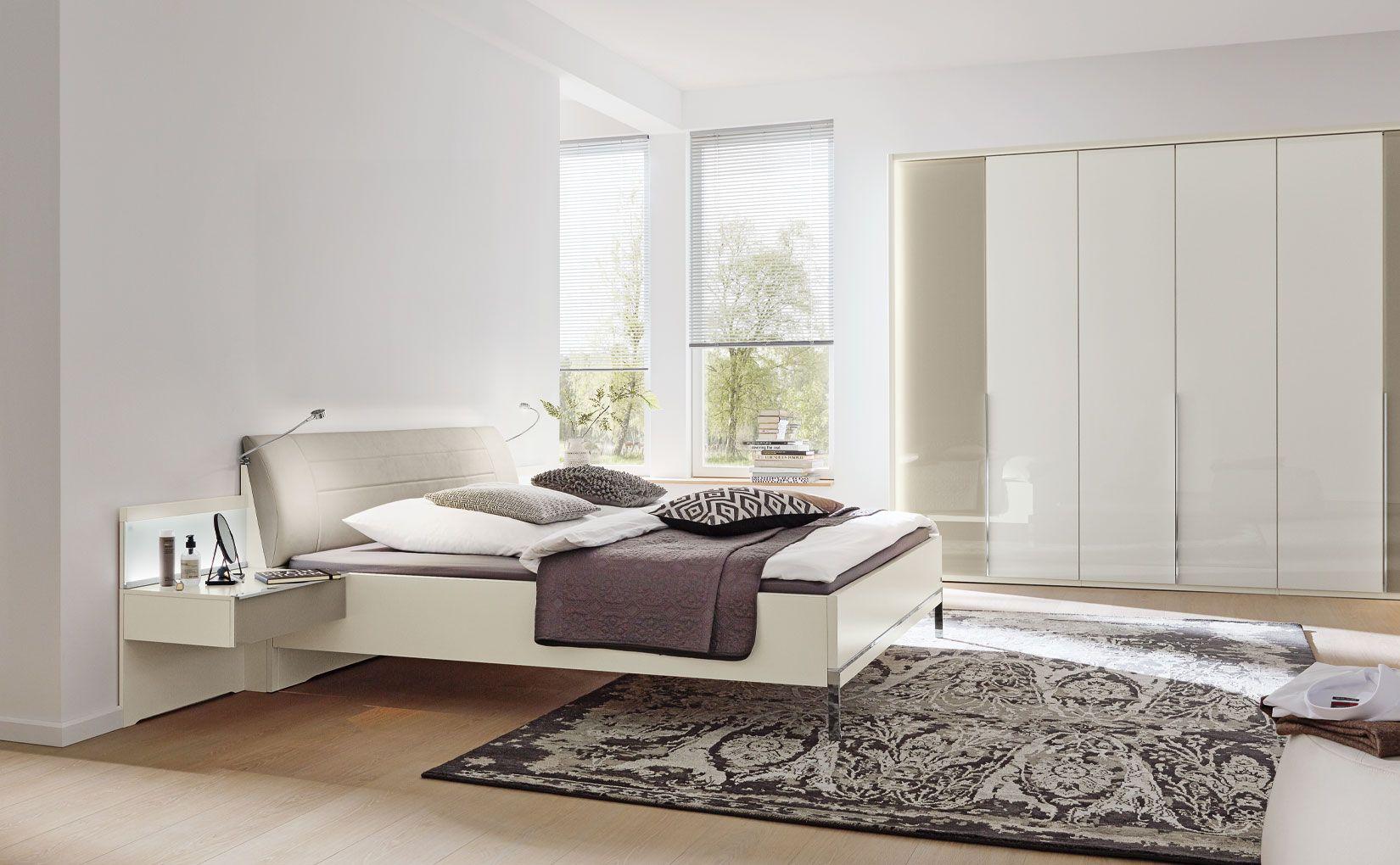 Schlafzimmer San Diego von Musterring mit hellem Polsterbett, integrierter Beleuchtung und großem Hochglanz-Kleiderschrank.